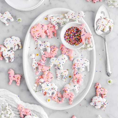 white and pink frosted animal cracker cookies on small white oval plate lined with parchment paper with small flowers and small bowl of rainbow sprinkles, more cookies around plate, with a white cheesecloth, stack of plates, a cake knife, and more flowers around on marble surface.