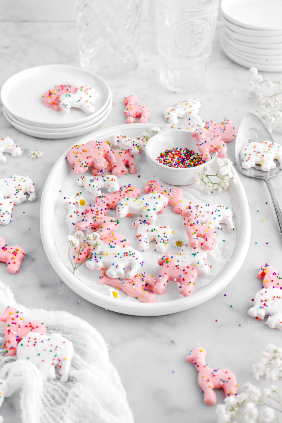 angled front shot of oval platter with pink and white frosted animal cracker cookies with more cookies around on marble surface with plates and empty glasses behind.