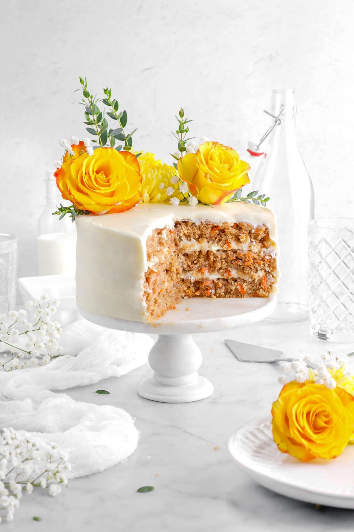 cut carrot cake on marble cake stand with yellow flowers and eucalyptus stems on top, with glasses behind, white flower around, a white cheese cloth beside, and a plate with yellow flowers in front.