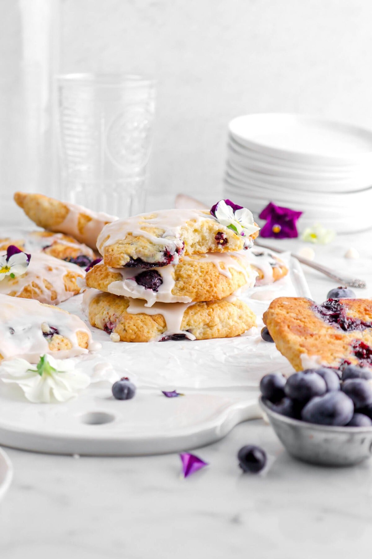 front shot of stacked scones with top scone missing a bite and a flower on top, with more scones around on white tray, with purple and white flowers, and blueberries scattered around on marble surface with stack of plates and empty glasses behind.