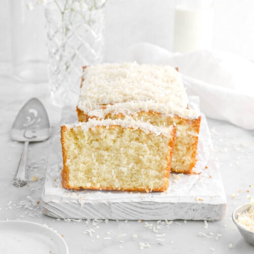 coconut pound cake with two slices leaning against loaf on white wood board with plates and a cake knife beside, a white napkin, white flowers, and glass of milk behind on marble surface.