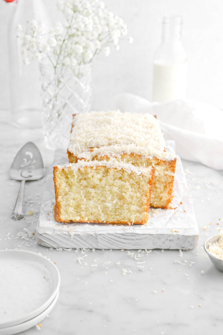 coconut pound cake with two slices leaning against loaf on white wood board with plates and a cake knife beside, a white napkin, white flowers, and glass of milk behind on marble surface.