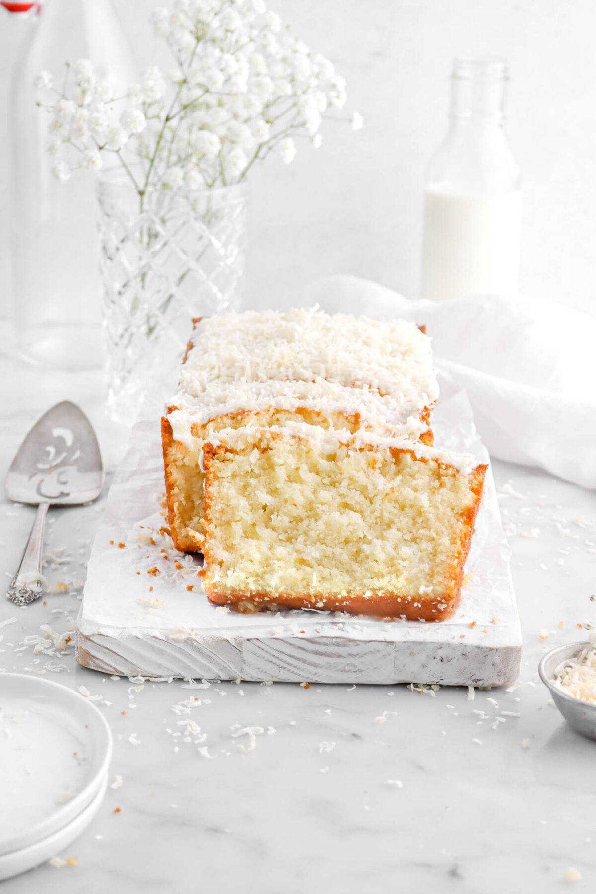 slice of coconut loaf cake leaning against loaf on white wood board with shredded coconut around, a cake knife beside, and white flowers behind with a glass of milk.