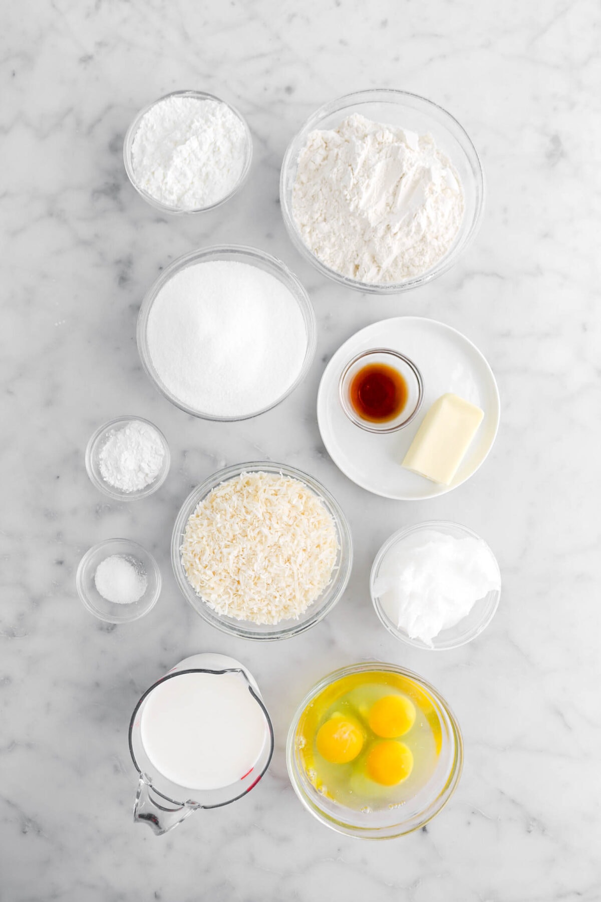corn starch, flour, sugar, vanilla, butter, baking powder, salt, shredded coconut, coconut oil, milk, and eggs on marble surface.