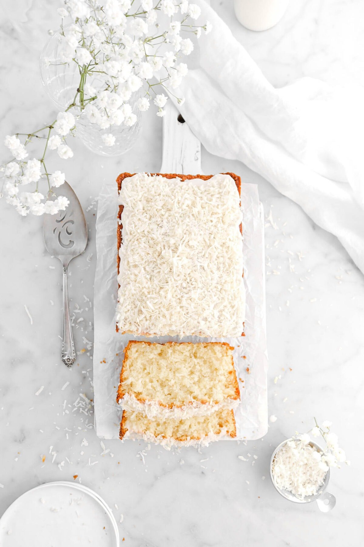 overhead shot of coconut pound cake with two slices laying in front with white flowers above, a cake knife and white napkin beside, and shredded coconut around.