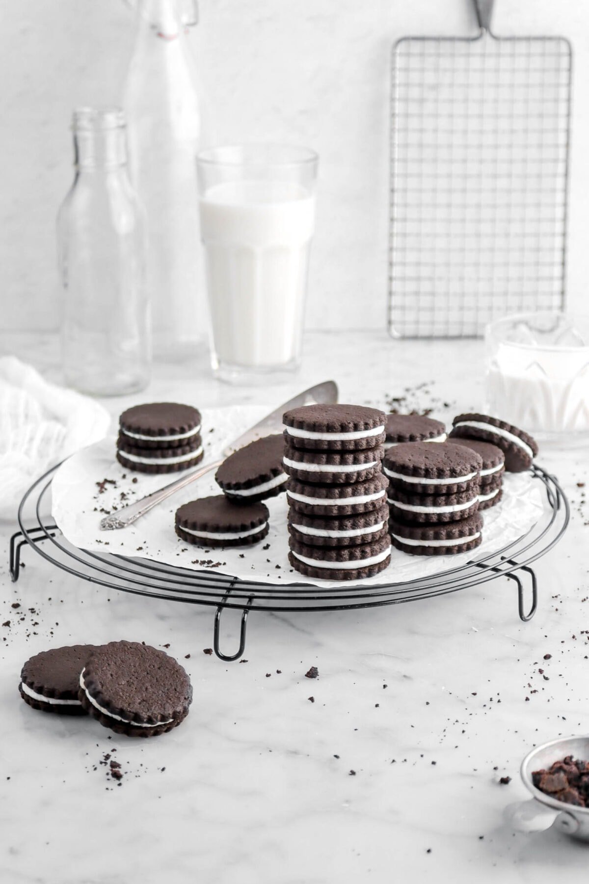 pulled back shot of stacked oreos on circle cooling rack with parchment paper and cake knife, with two glasses of milk behind, and cooling rack.