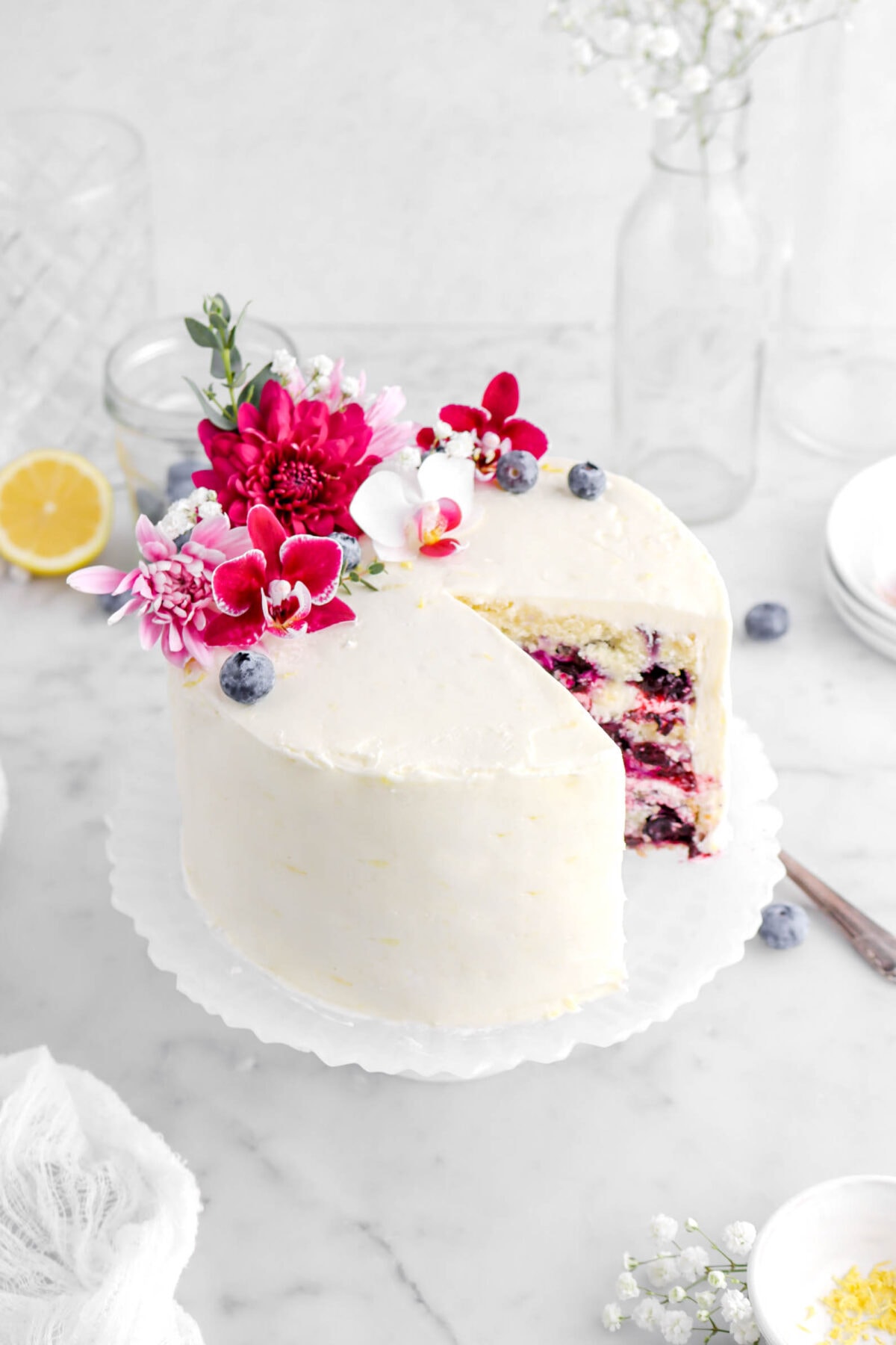 angled close up of lemon blueberry layer cake decorated with flowers on top with lemon half and empty glass behind on marble surface.