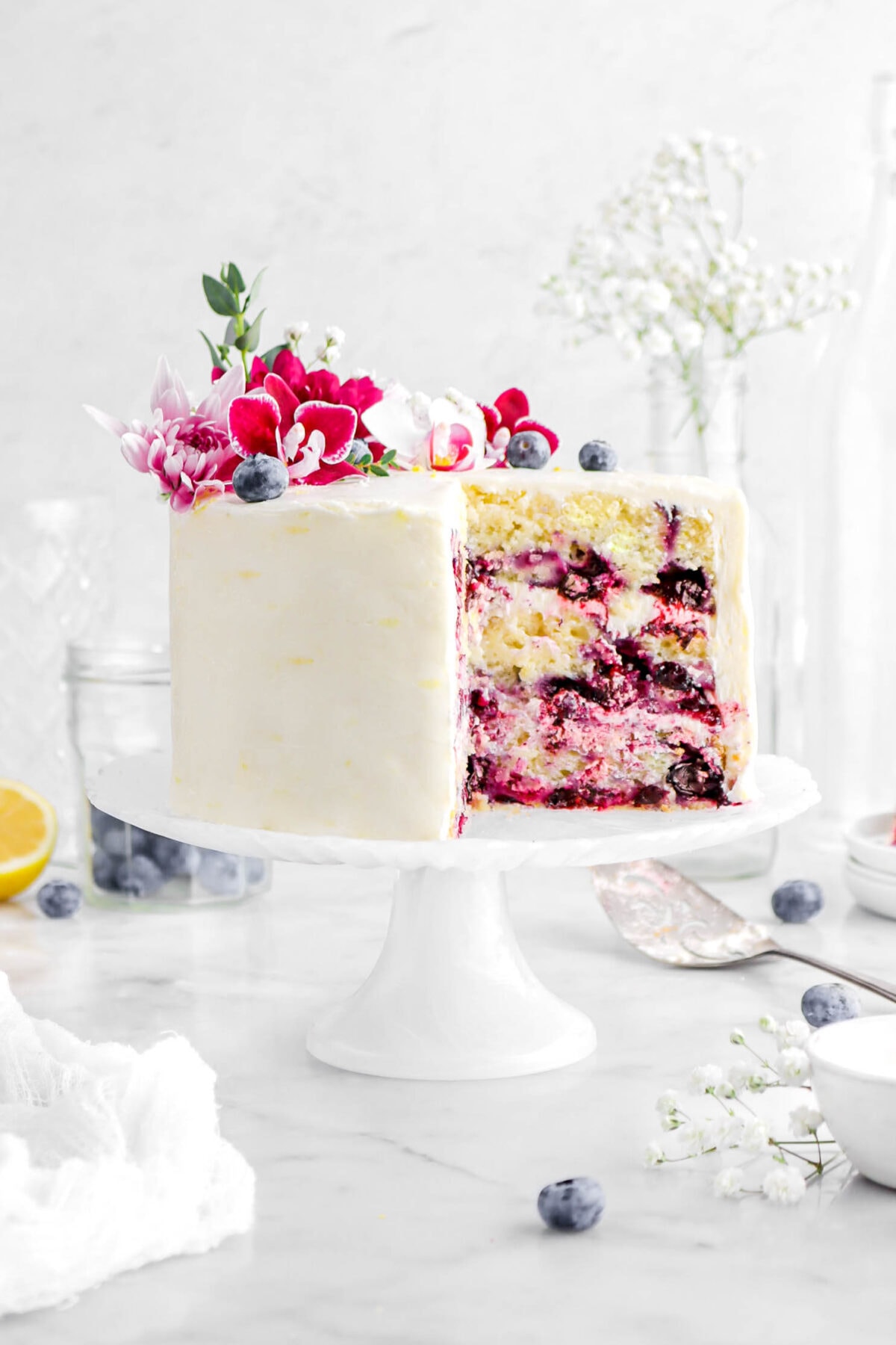 close up of cut lemon blueberry cake with blueberries and flowers on top on a white cake stand with blueberries and white flowers around on marble surface.
