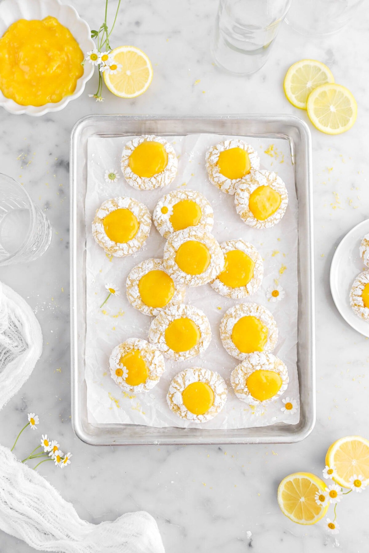 thirteen lemon curd thumbprint cookies on small lined sheet pan with chamomile flowers on some cookies, with lemon halves, a bowl of lemon curd, an empty glass, and more chamomile flowers around on marble surface.
