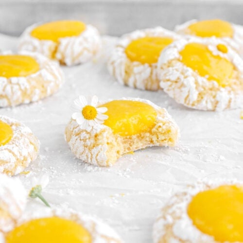 front shot of lemon curd thumbprint cookie with bite missing, with more cookies around on lined sheet pan with lemon halves and more flowers behind.