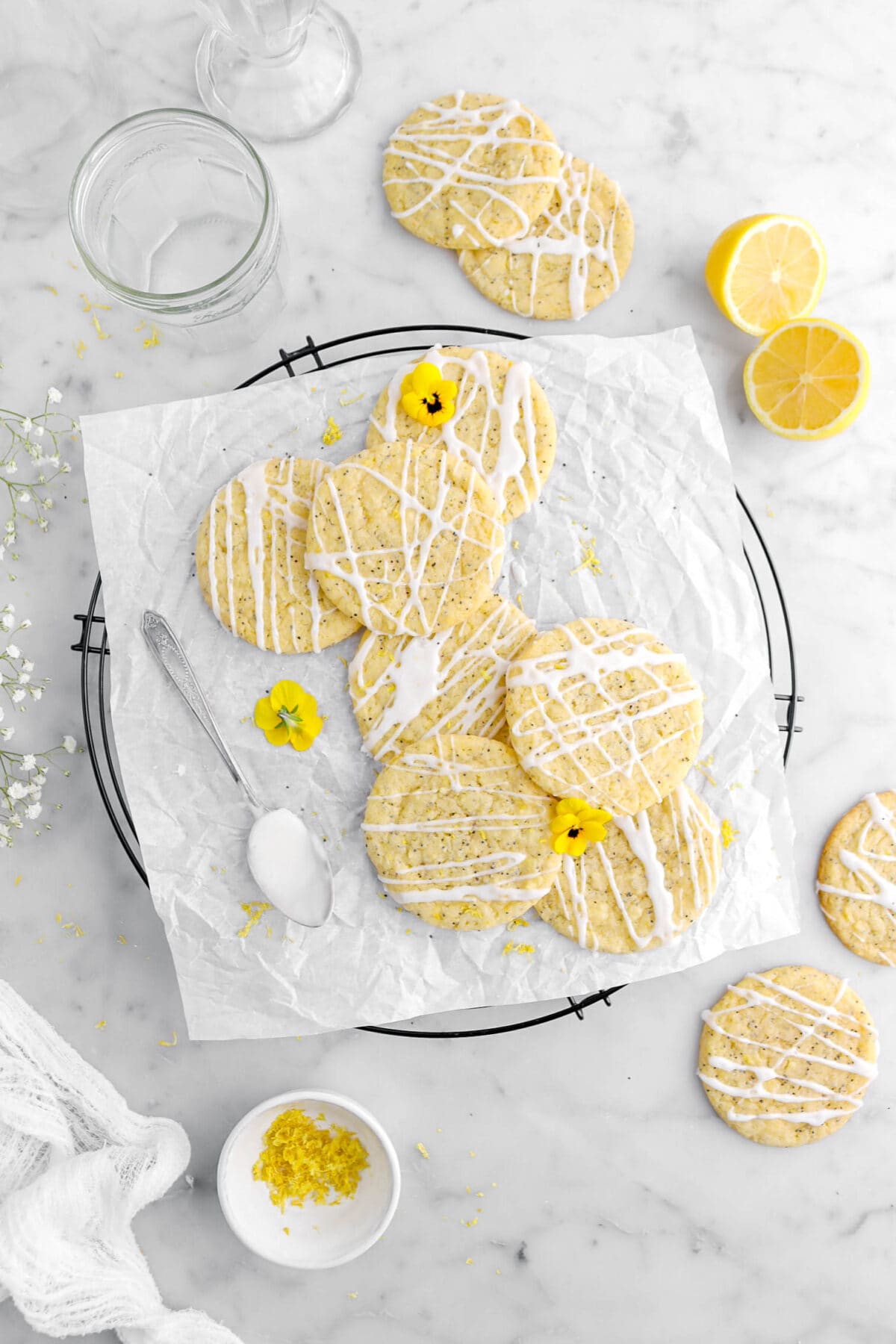 overhead shot of seven lemon poppy seed cookies on parchment lined cooling rack, with four more cookies and lemon halves around on marble surface with bowl of lemon zest.
