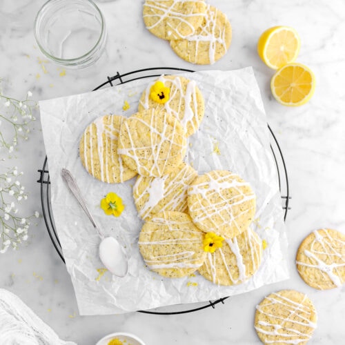 pulled back overhead shot of lemon poppy seed cookies on parchment lined cooling rack with yellow flowers on top and spoon of icing beside, with four more cookies around on marble surface, with two lemon halves and empty glasses above.