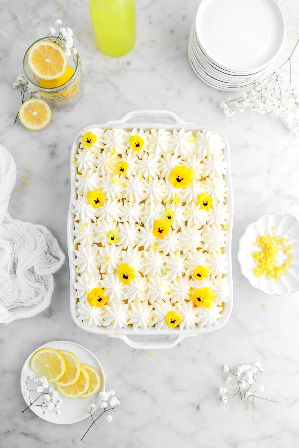 pulled back overhead shot of lemon tiramisu with yellow pansies on top with bowl of lemon zest beside, a plate of lemon slices, stack of white places, and glass of limoncello around on marble surface.