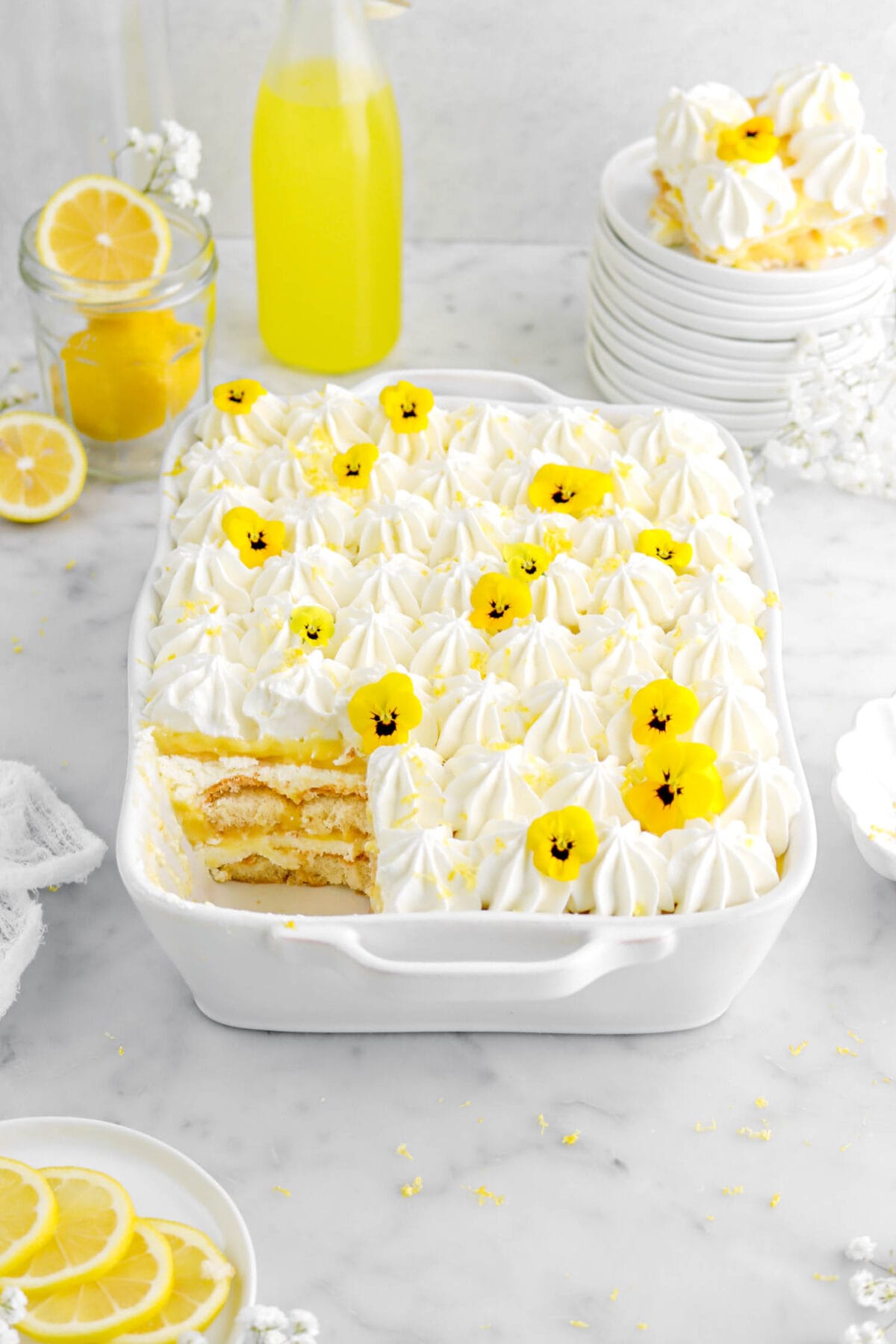 angled shot of lemon tiramisu with left corner missing a slice in white casserole with lemons and white flowers around, stack of white plates with slice tiramisu behind.