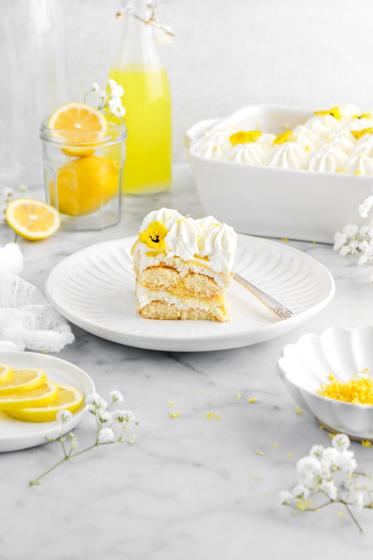 pulled back shot of lemon tiramisu slice on white plate with fork beside, white flowers around, lemons around, and casserole of tiramisu behind.
