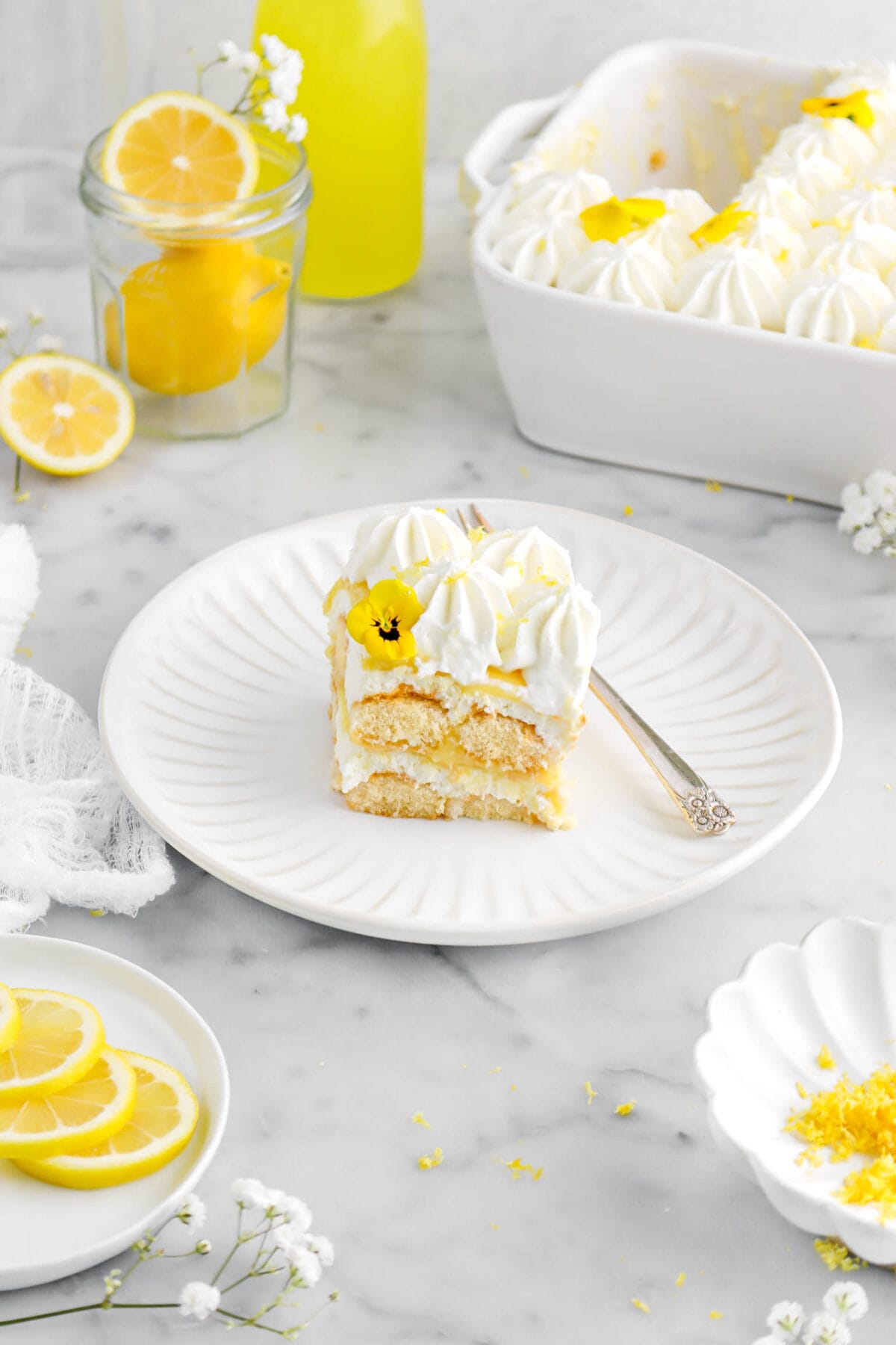 angled shot of lemon tiramisu on white plate with fork beside with white flowers around, lemons, and white casserole with tiramisu behind.