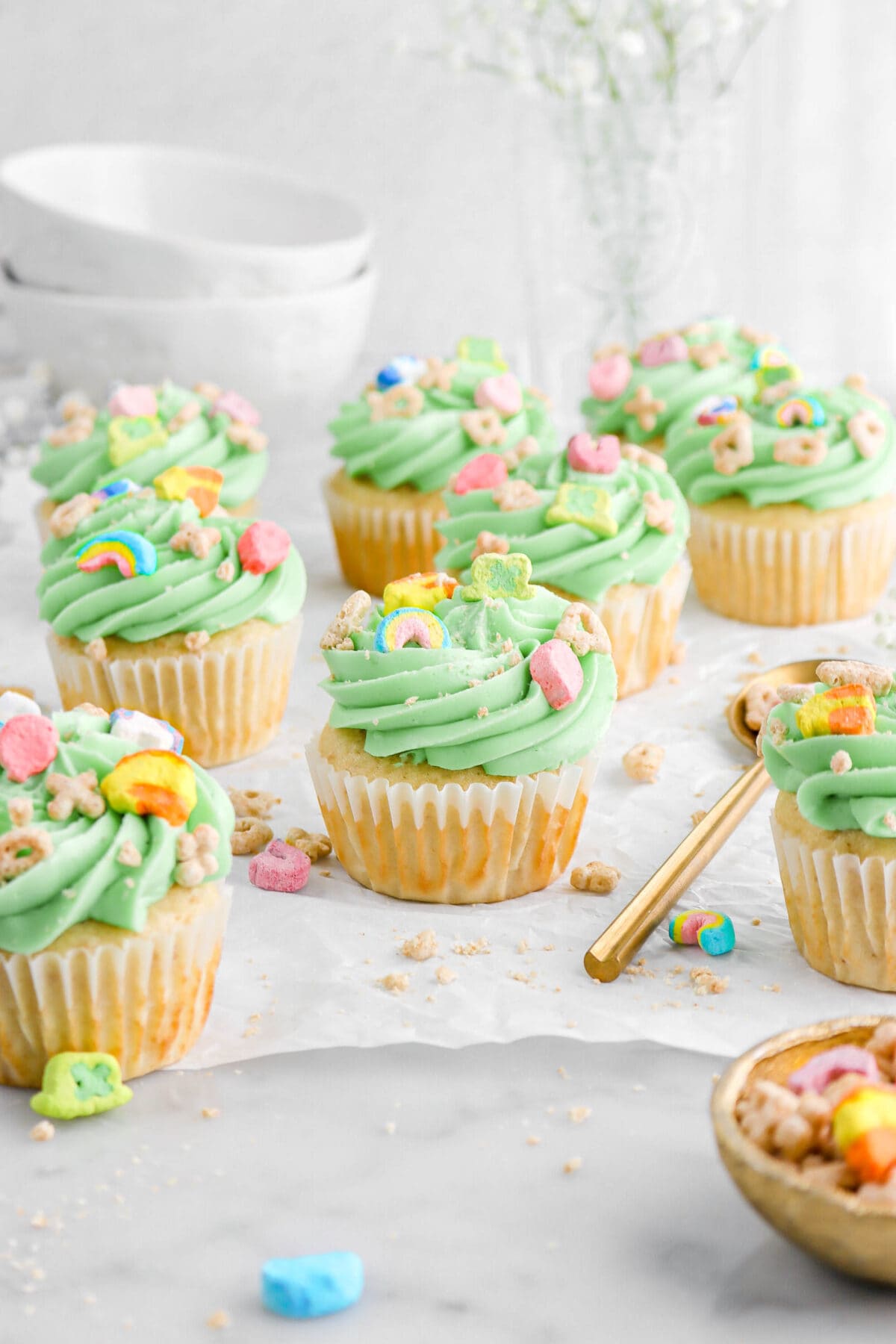 nine lucky charms cupcakes on parchment paper with cereal scattered around, a gold bowl and gold spoon beside cupcakes with white flowers behind, as well as stack of mugs.