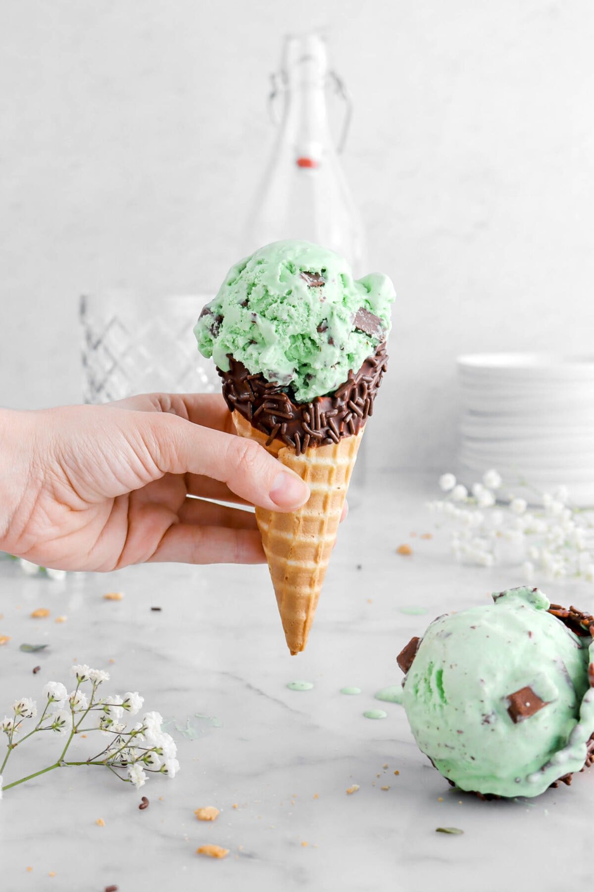 hand holding cone of mint chip ice cream with another cone of ice cream beside and white flowers around.