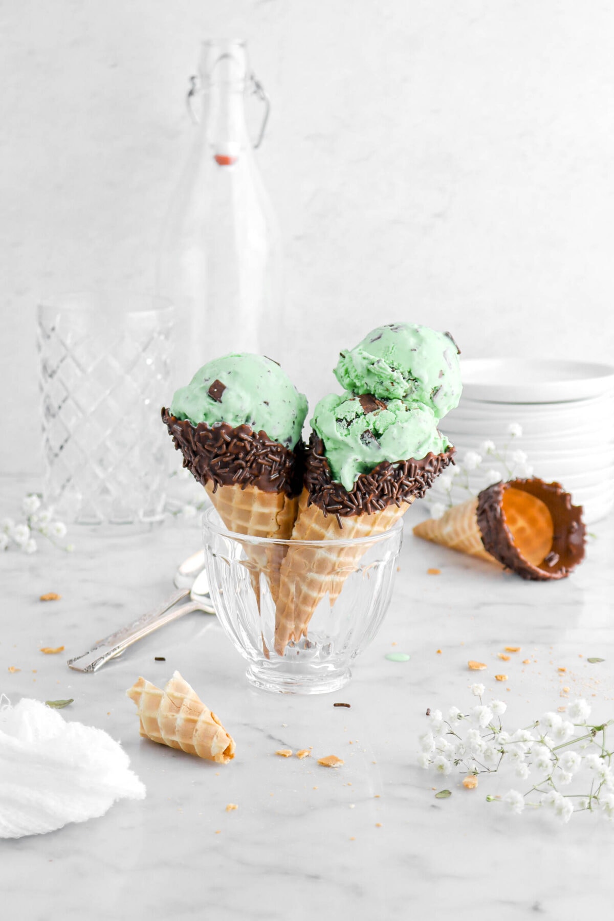 two cones of mint chip ice cream in glass with one cone having two scoops of ice cream with white flowers and crumbs around.