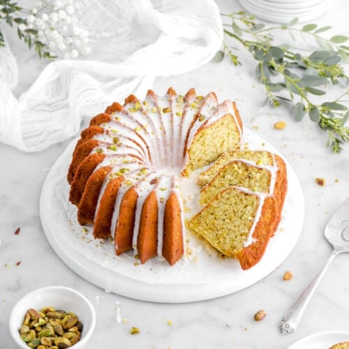angled shot of pistachio cake on upside down plate with three slices laying beside on plate with cake knife and a small plate of cake slices beside, eucalyptus branches, white flowers, and white cheesecloth behind.
