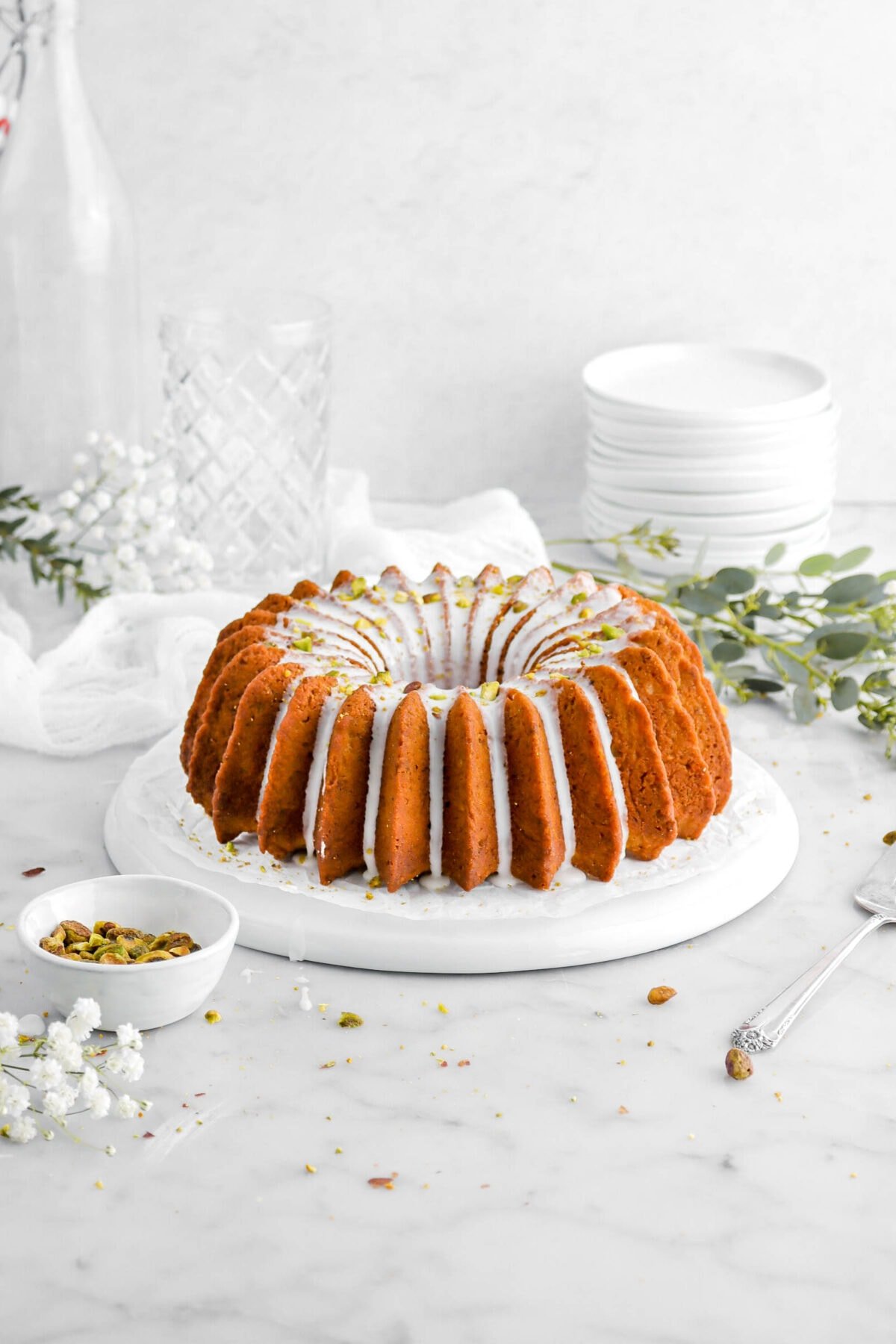 bundt cake on upside down white palte with icing on top and pistachios, with eucalyptus leaves behind, stack of plates, empty glasses, and white flowers.