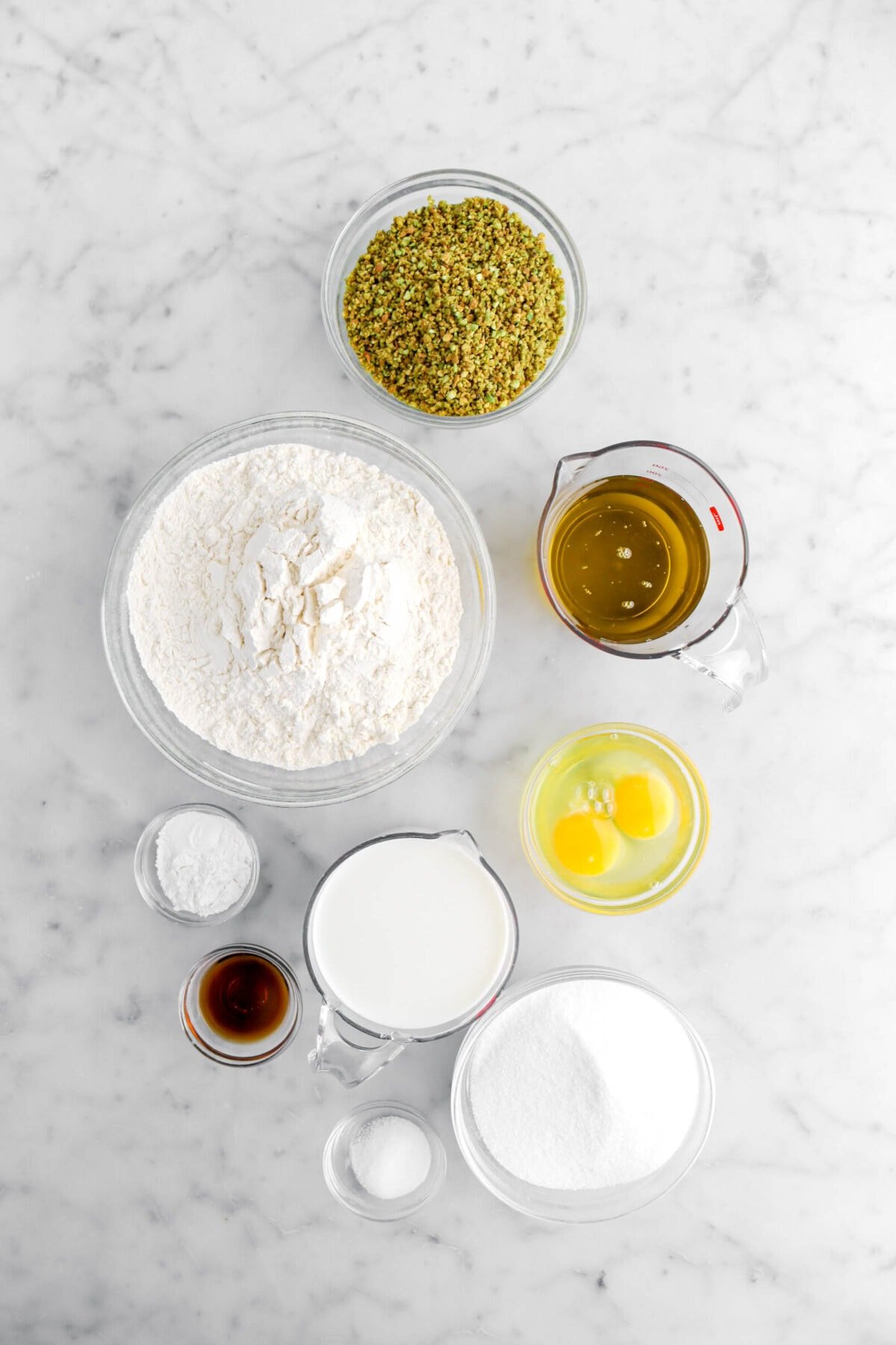 ground pistachios, olive oil, flour, eggs, baking powder, vanilla, milk, sugar, and salt on marble surface.