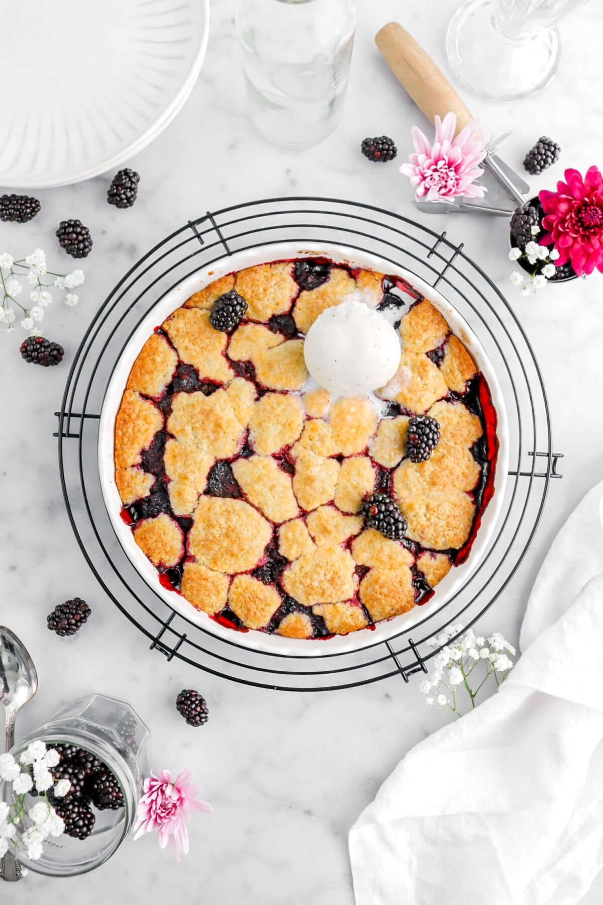 blackberry cobbler in round white pan on a round black wire cooling rack with a scoop of ice cream and three fresh blackberries on top of cobbler, with more berries and flowers surrounding dish on marble surface with white napkin and white plate beside.