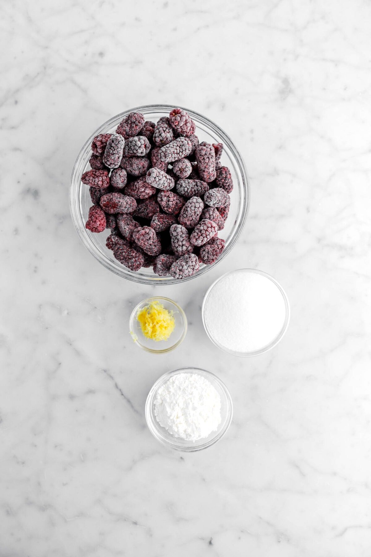 frozen blackberries, lemon zest, sugar, and corn starch in glass bowls on marble surface.