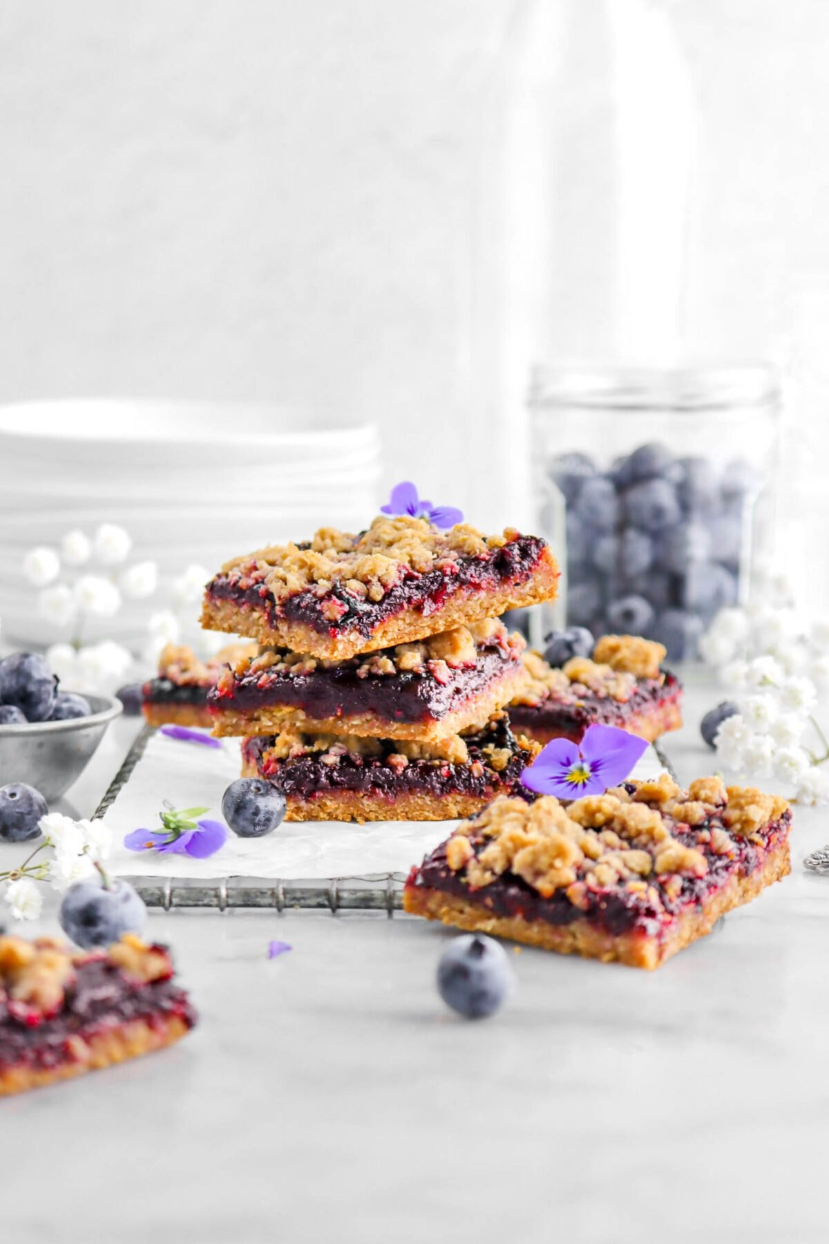 three stacked blueberry crumble bars with more around, with blueberries, white flowers, and purple flowers around on marble surface.
