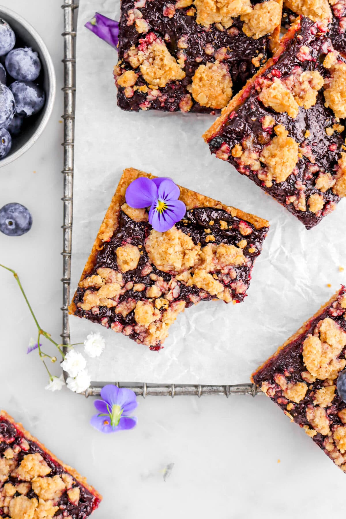 overhead shot of blueberry crumble bar on parchment paper lined tray with bite missing and more crumble bars around, a bowl of blueberries, and white flowers beside.