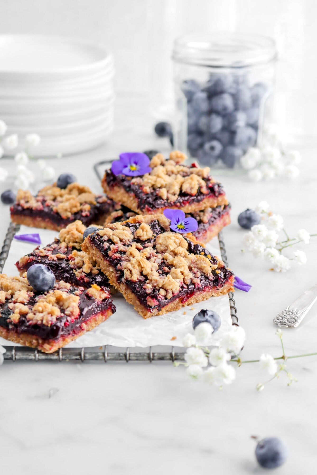 six blueberry crumble bars on parchment paper lined wire tray with blueberries and flowers around on marble surface.