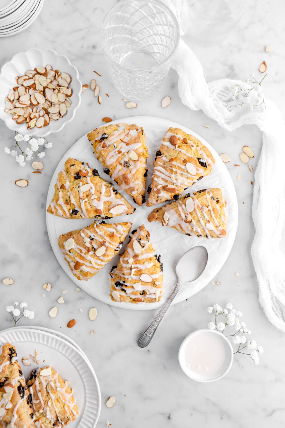 overhead shot of cherry almond scones on upside down white plate with white flowers and almond around on marble surface.