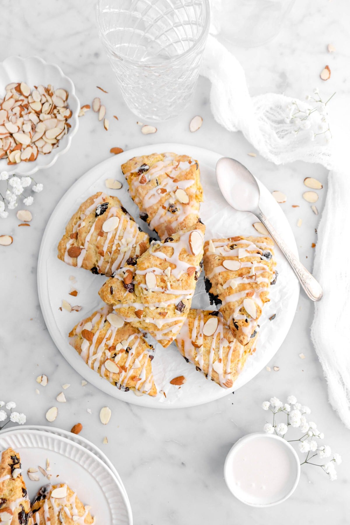scones piled on upside down white plate with spoon beside, white flowers, almonds, and a cheese cloth around on marble surface.