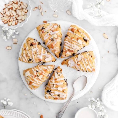 six scones on upside down plate with spoon beside and sliced almonds in bowl with white flowers and white cheese cloth around on marble surface.