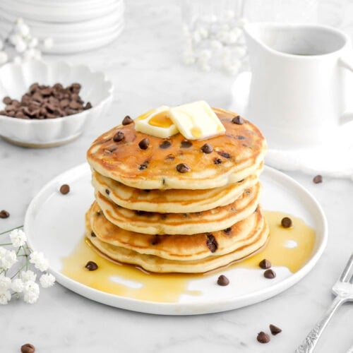 five stacked chocolate chip pancakes with butter and maple syrup on top of the top pancake, with bowl of chocolate chips behind, a ceramic creamer, and flowers behind.