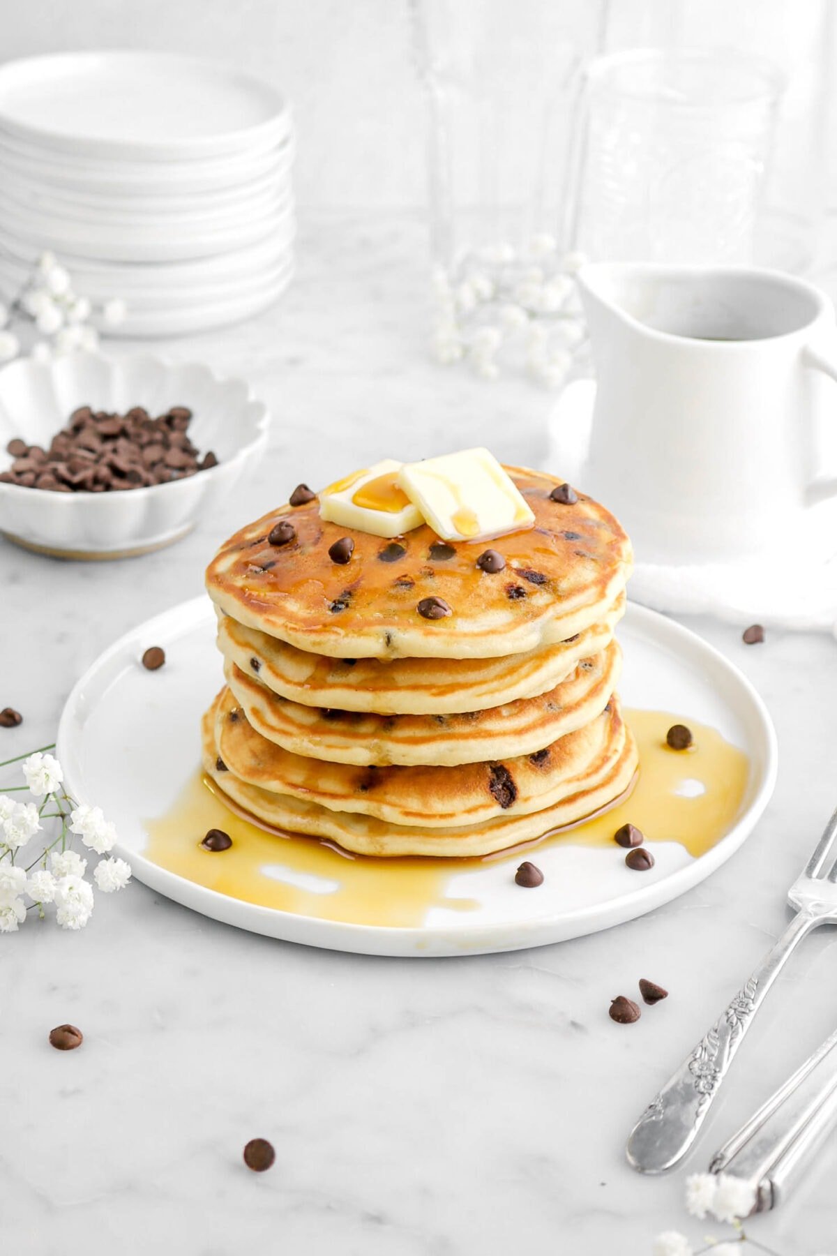 five stacked chocolate chip pancakes with butter and maple syrup on top of the top pancake, with bowl of chocolate chips behind, a ceramic creamer, and flowers behind.