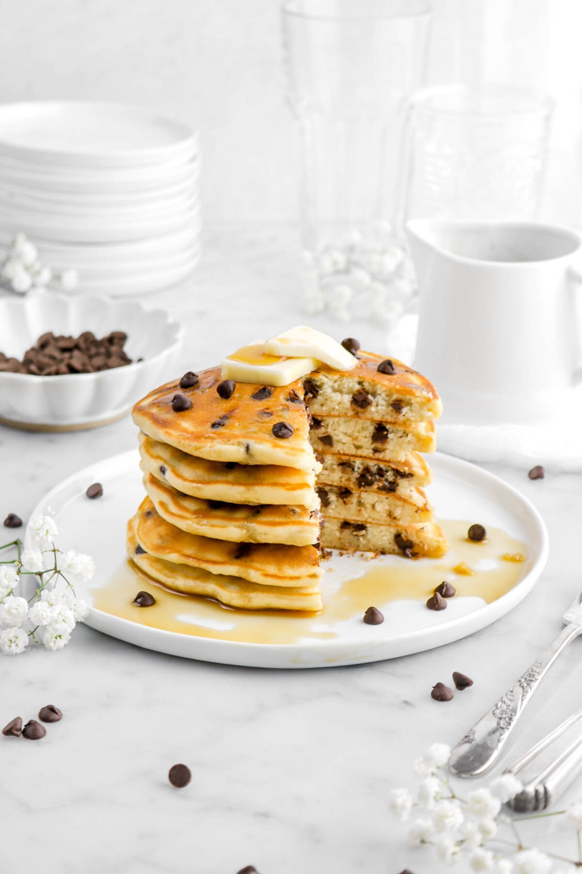 cut stacked pancakes on white plate with butter and chocolate chips on top, with white flowers, and more chocolate chips around.