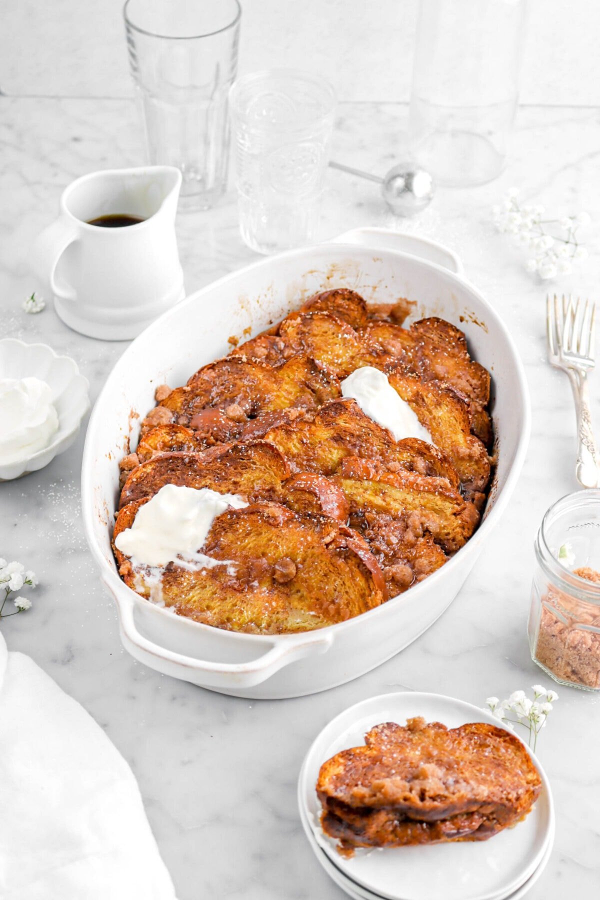 french toast in oval casserole with two dollops of whipped cream on top, with a slice of french toast on stack of white plates, with forks, white flowers, and empty glasses behind.