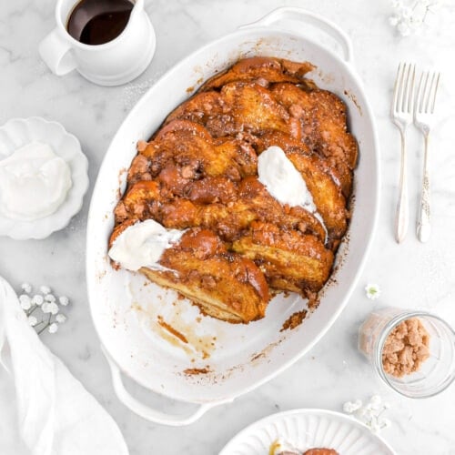 overhead shot of french toast casserole with slices on white plate beside, with jar of streusel, forks, bowl of whipped cream, and cup of maple syrup beside.
