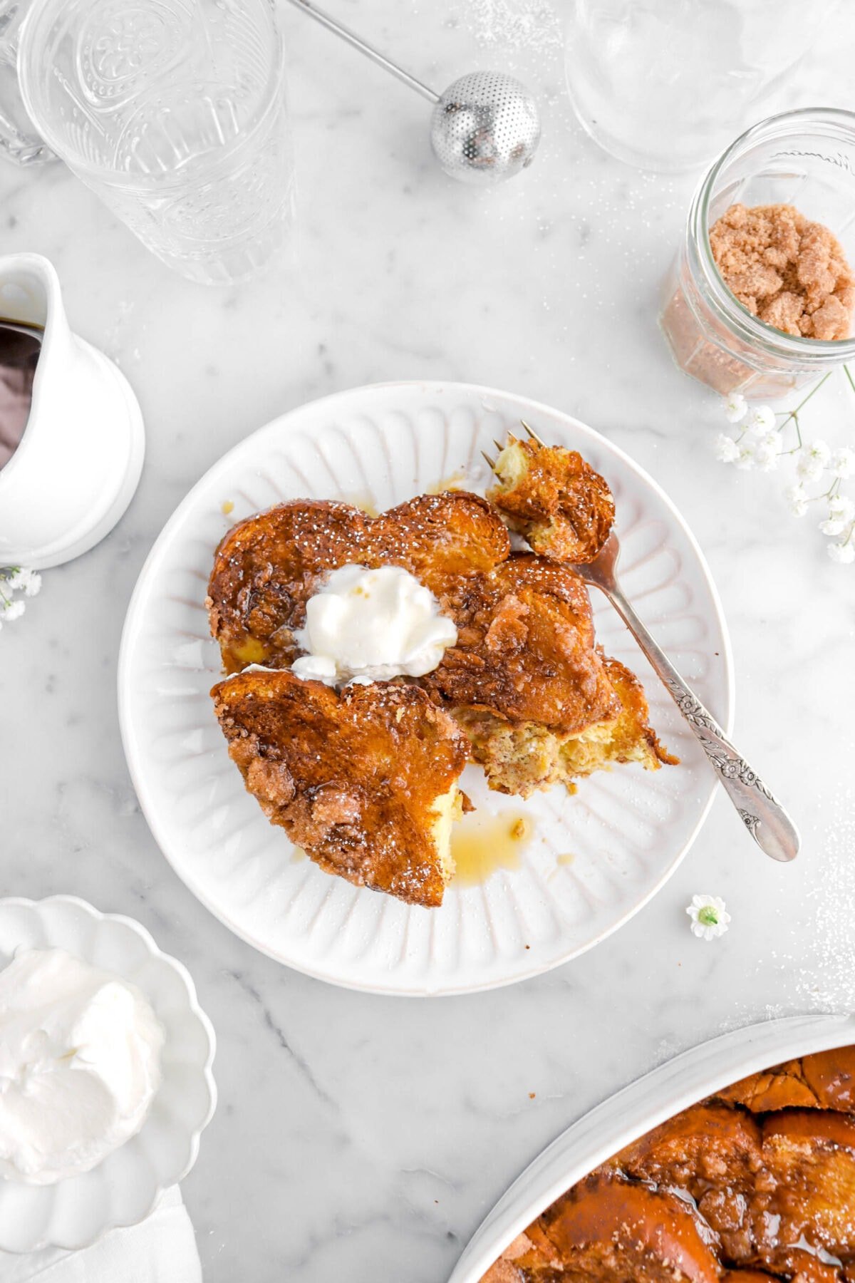overhead shot of french toast with forkful of french toast beside, with jar of streusel, white flowers, bowl of whipped cream, and casserole around.