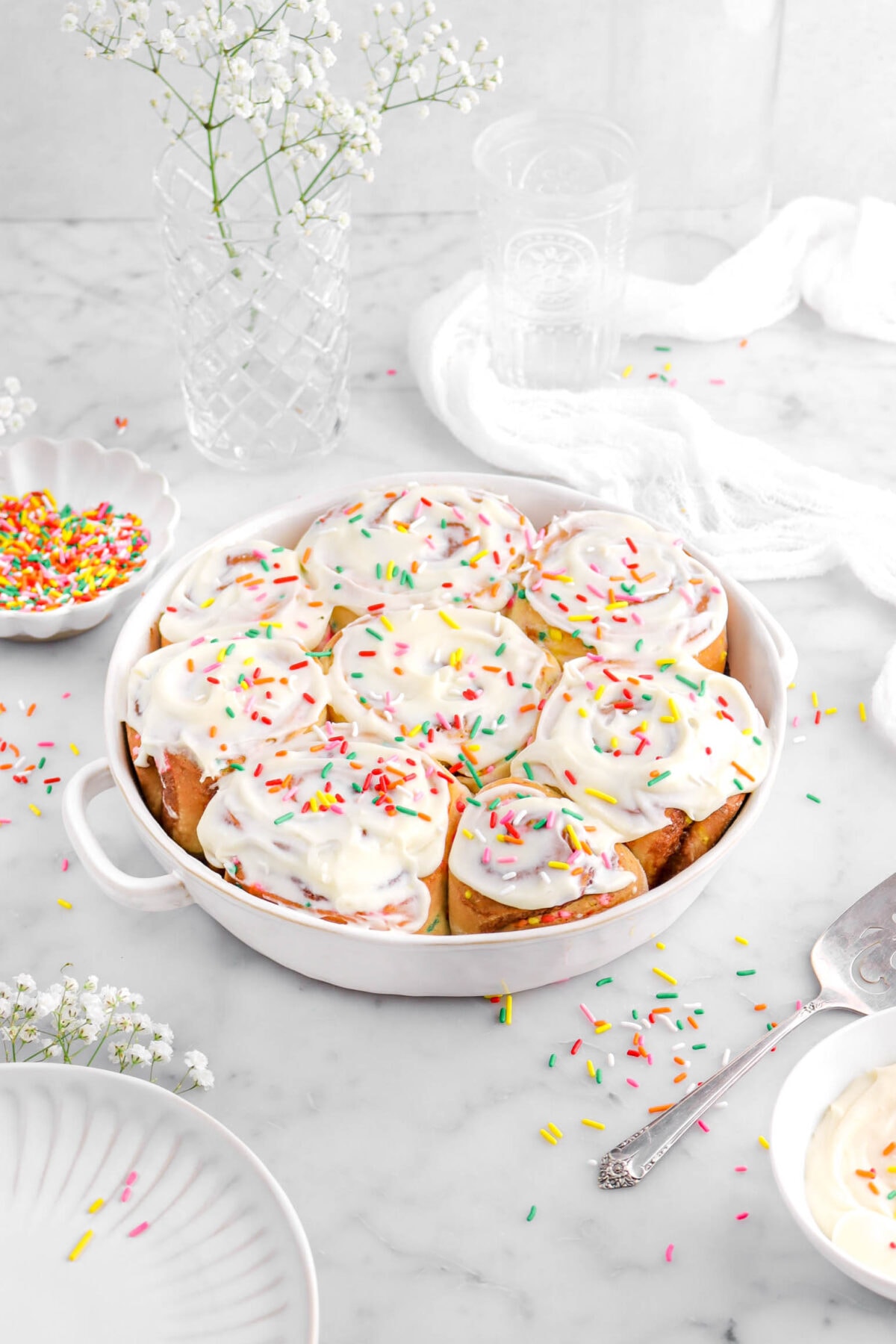 angled image of eight cinnamon rolls in round white cassserole with frosting and sprinkles on top, with sprinkles around, a cake knife beside with bowl of frosting and bowl of sprinkles, a white cheese cloth, a white plate, with white flowers around on marble surface.