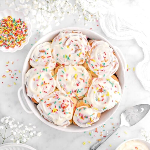 overhead shot of eight cinnamon rolls in white casserole with cream cheese frosting and rainbow sprinkles on top, with cake knife, bowl of frosting, bowl of sprinkles, and white flowers around.