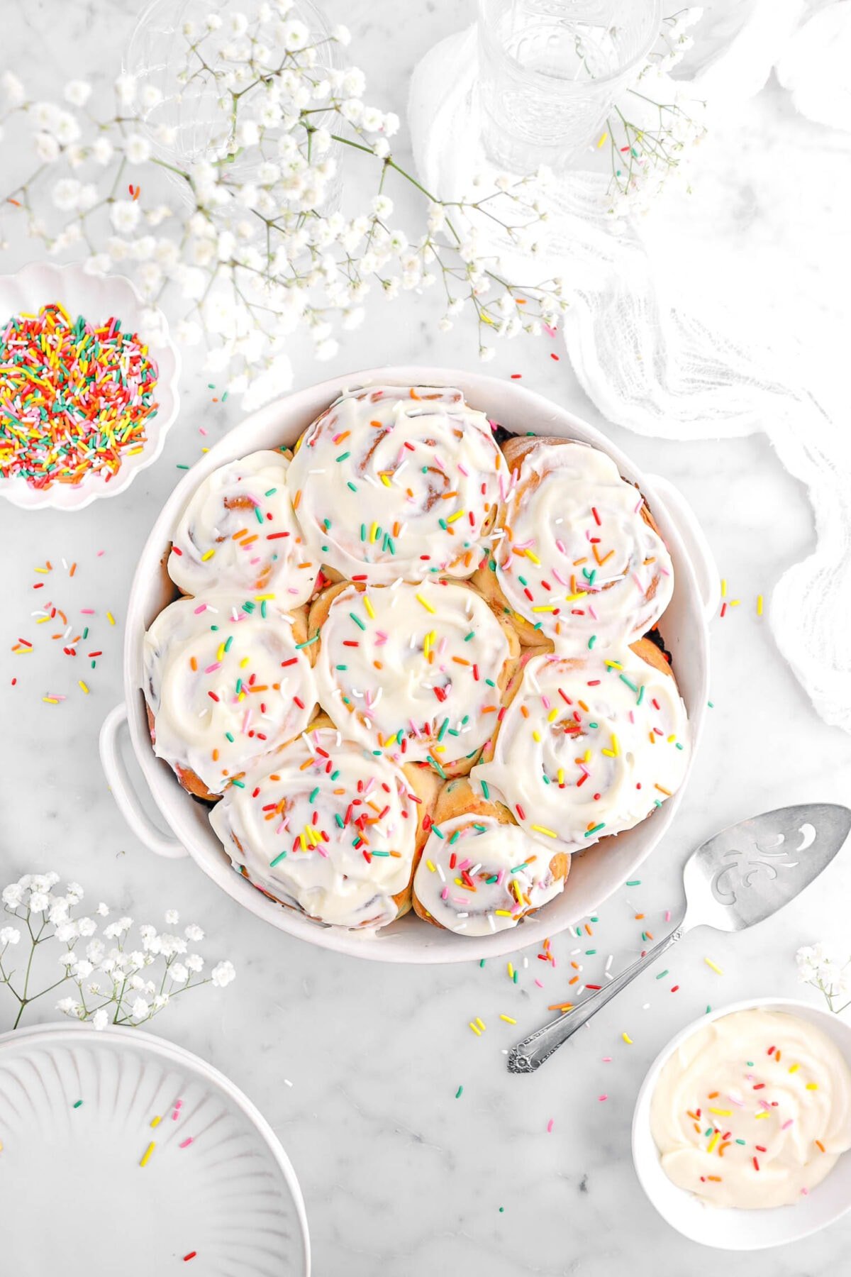 overhead shot of eight cinnamon rolls in white casserole with cream cheese frosting and rainbow sprinkles on top, with cake knife, bowl of frosting, bowl of sprinkles, and white flowers around.