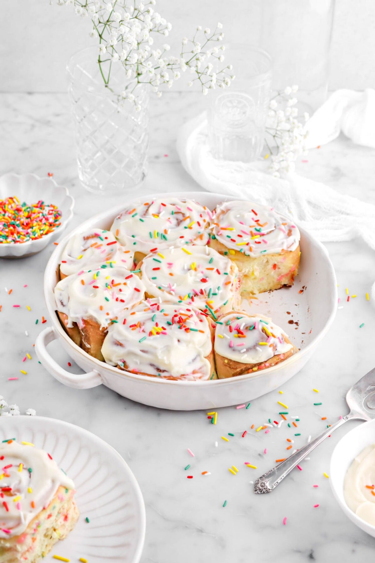 angled shot of frosted funfetti cinnamon rolls in round casserole with one cinnamon roll missing, with rainbow sprinkles around, a white cheese cloth, glasses, and white flowers behind on marble surface.