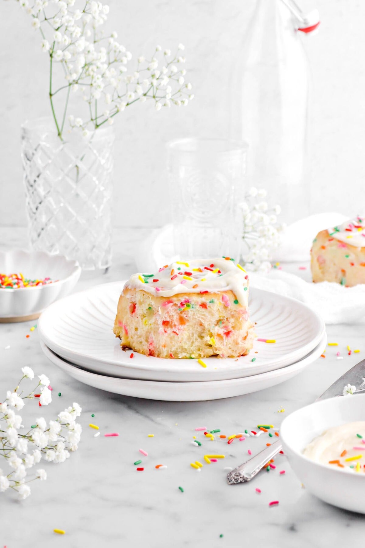 frosted funfetti cinnamon roll on stack of two plates with sprinkles and white flowers around, a white cheese cloth, empty glasses, and another cinnamon roll behind.