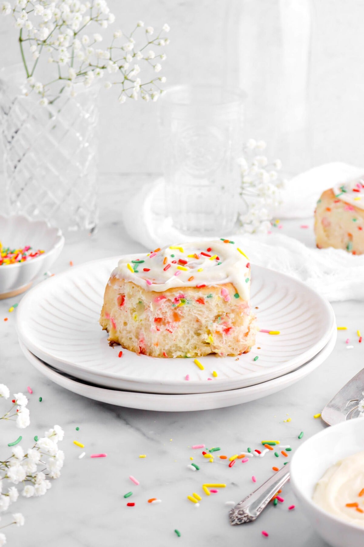 frosted cinnamon roll on two stacked white plates with empty glasses, a bowl of rainbow sprinkles, and another cinnamon roll behind, with white flowers and rainbow sprinkles around on marble surfce.