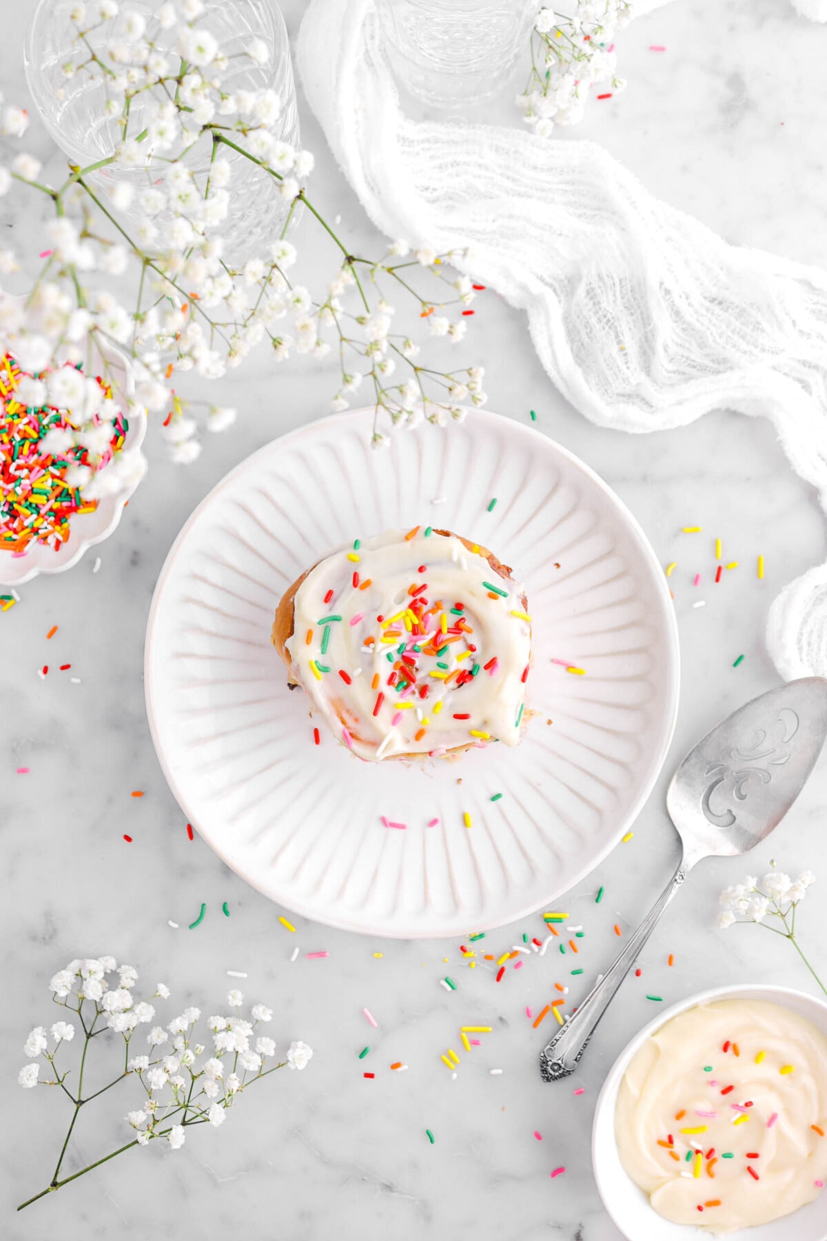 overhead shot of of frosted cinnamon roll with sprinkles on top on white plate with cake knife beside, white flowers, and white cheesecloth around.
