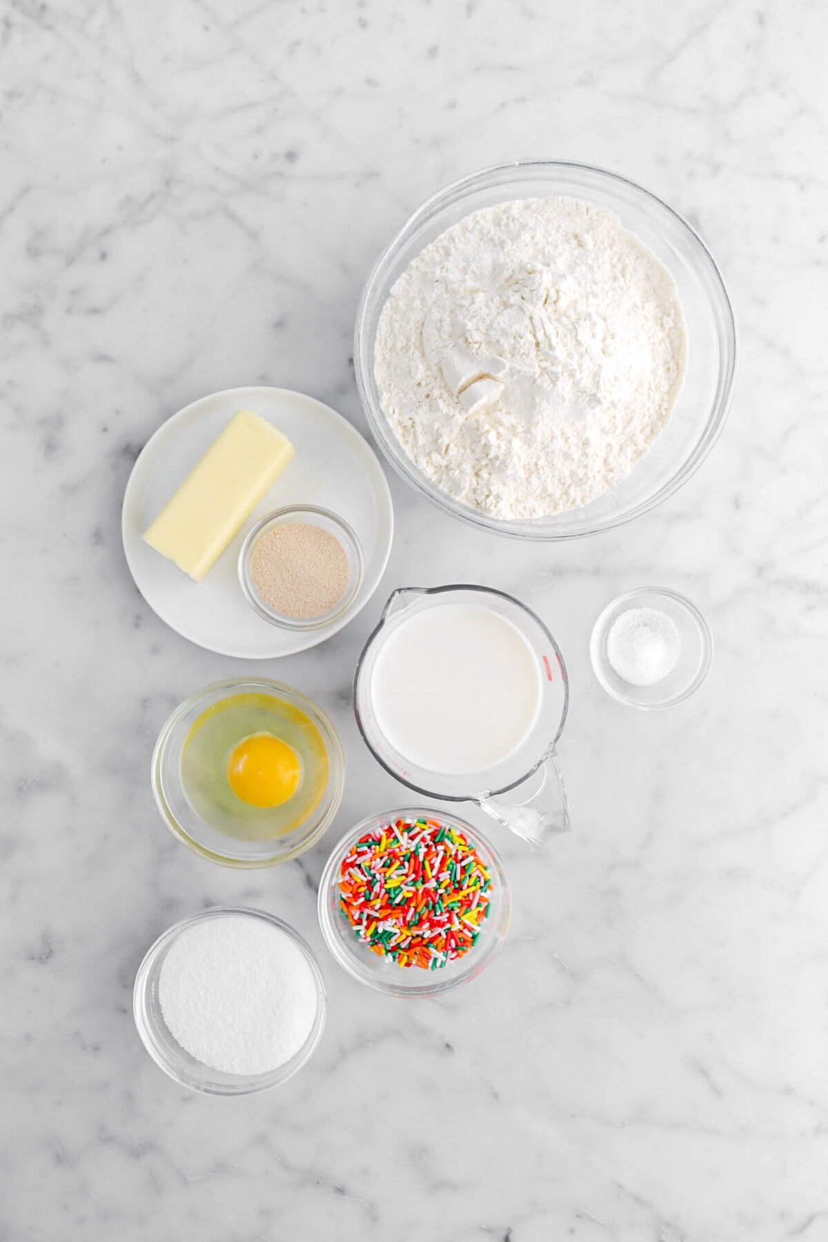 flour, butter, yeast, milk, salt, egg, rainbow sprinkles, and sugar on marble surface.