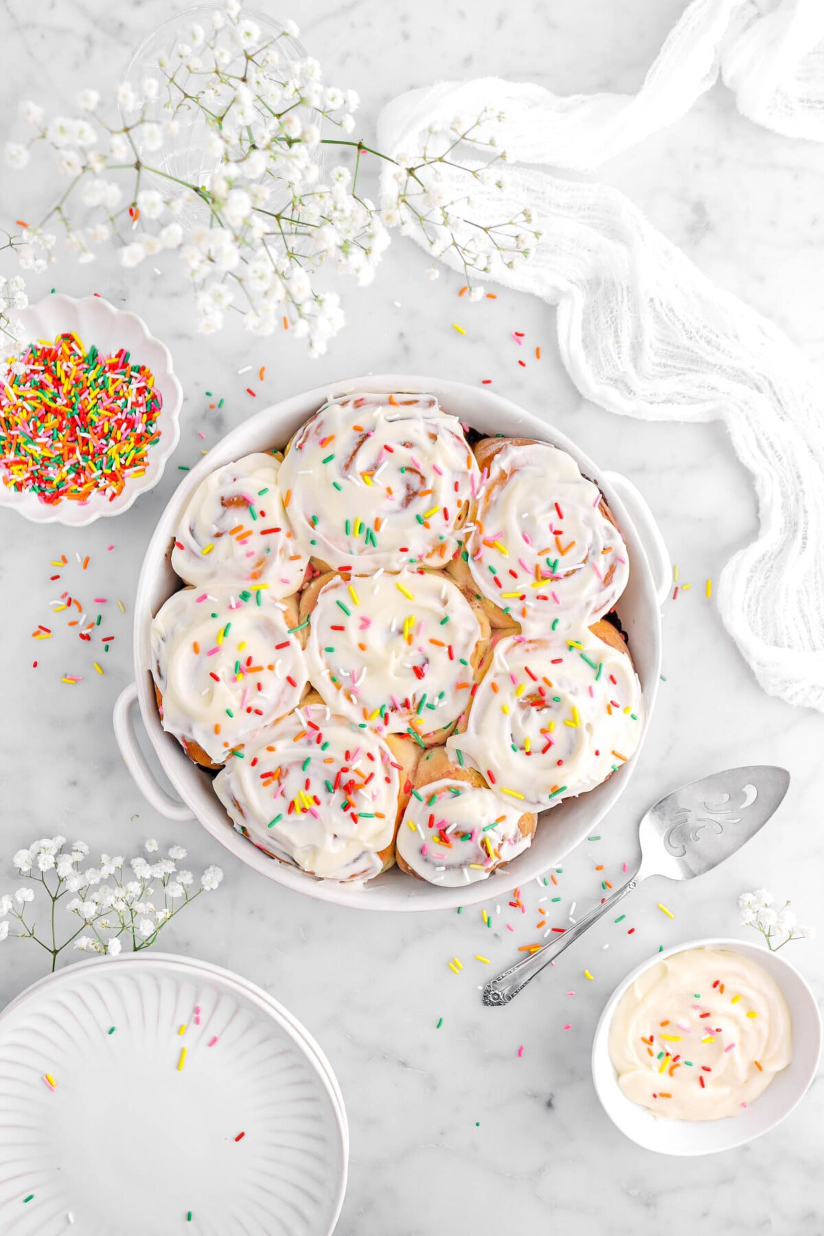 frosted cinnamon rolls with rainbow sprinkles on top in white casserole with a bowl of frosting and a bowl of sprinkles beside, white flowers, stack of plates, and a white cheesecloth around on marble surface.