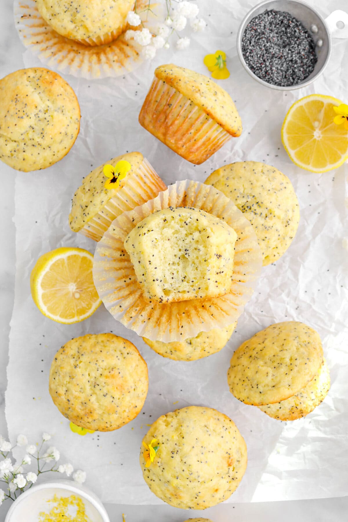 lemon poppy seed muffin on cupcake paper on top of more muffins with a bite missing, two lemon halves, yellow pansies, and white flowers around.